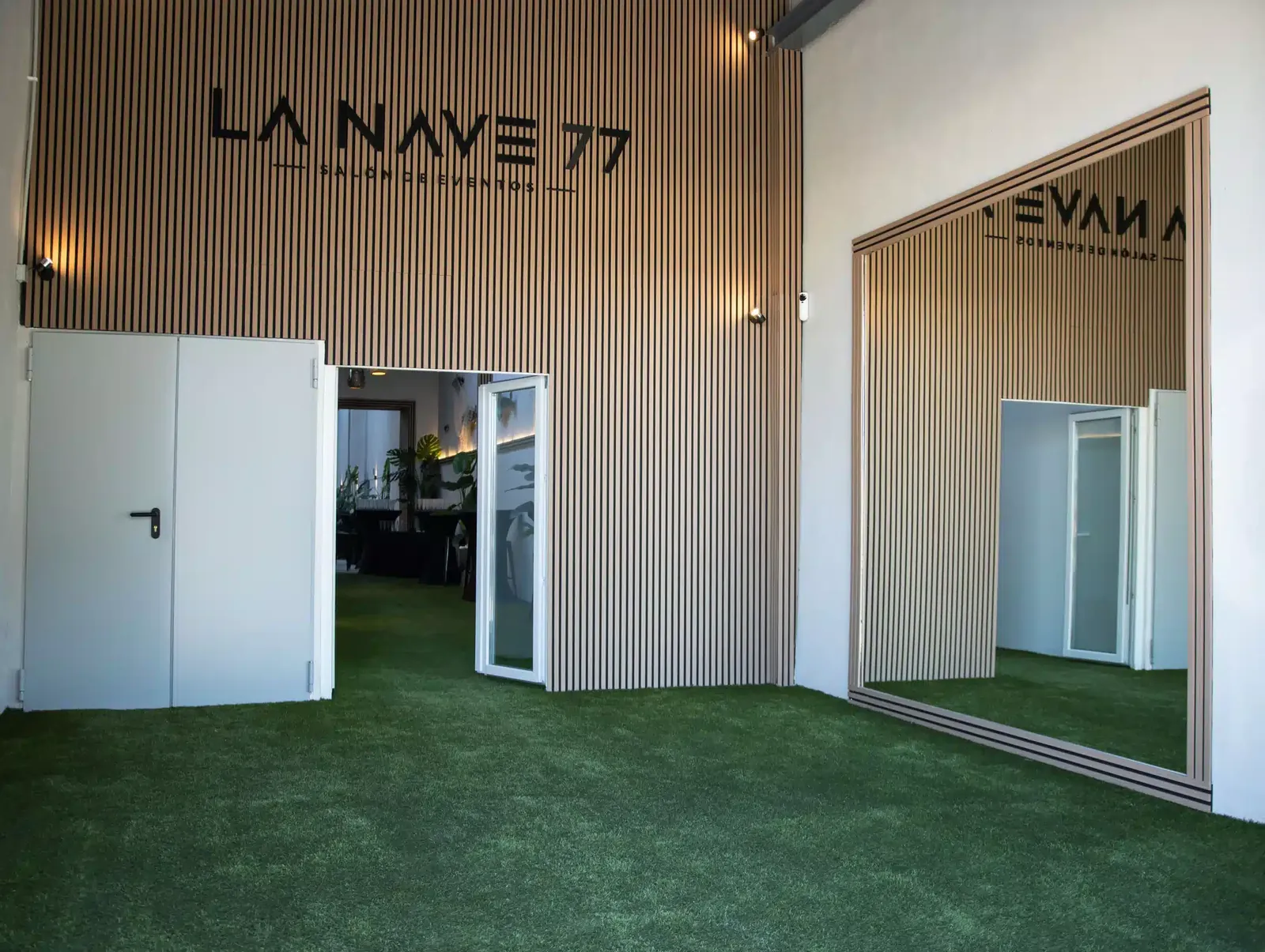 Recepción de LA NAVE 77 con el logotipo sobre pared de listones de madera y césped artificial