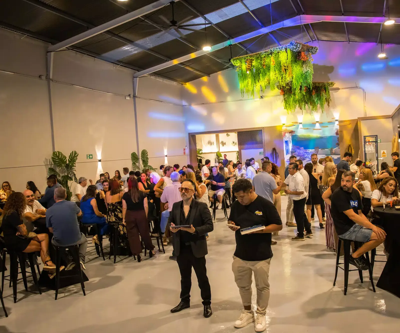 Salón de LA NAVE 77 en Málaga lleno de invitados durante la inauguración, con luces de colores y jardines colgantes